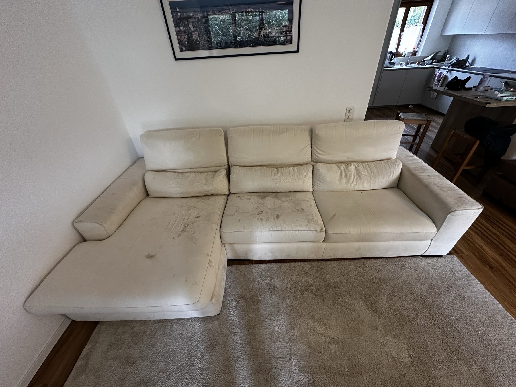 Divano angolare in tessuto color panna con macchie scure diffuse su sedute e chaise longue
